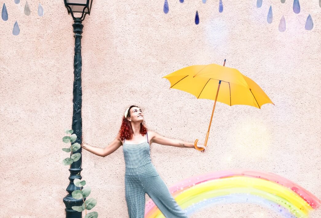 Creative photo of Julia holding a lamppost and an umbrella, with rainy clouds and a rainbow. Watercolour style.