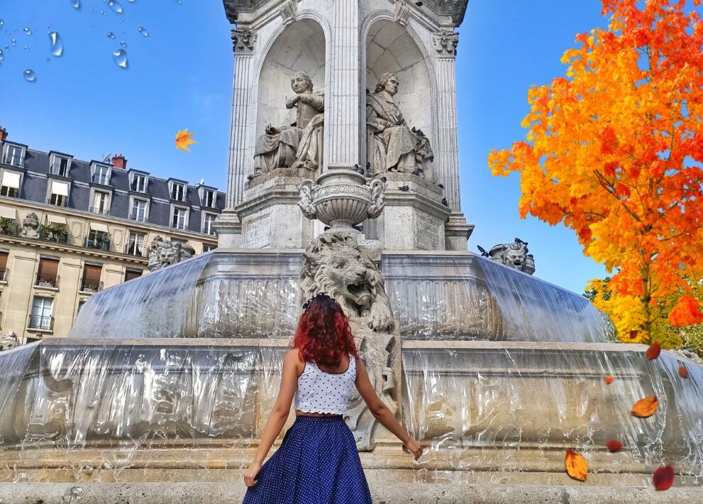 Picture of a girl standing if front of a big fountain in Paris, France. The sky is blue and there is a tree with orange autumn leaves in the background.