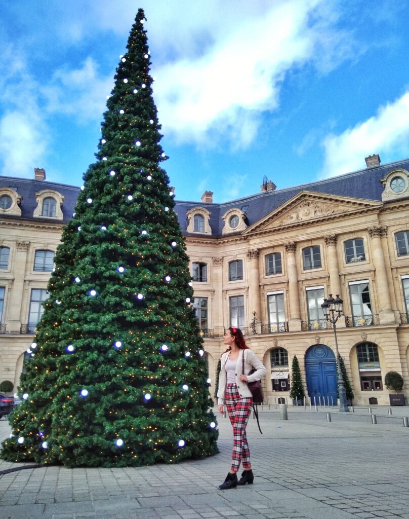 Plâce Vendôme in Paris, with a big Christmas tree in the foreground and girl dressed in Christmas colours posing near the tree.