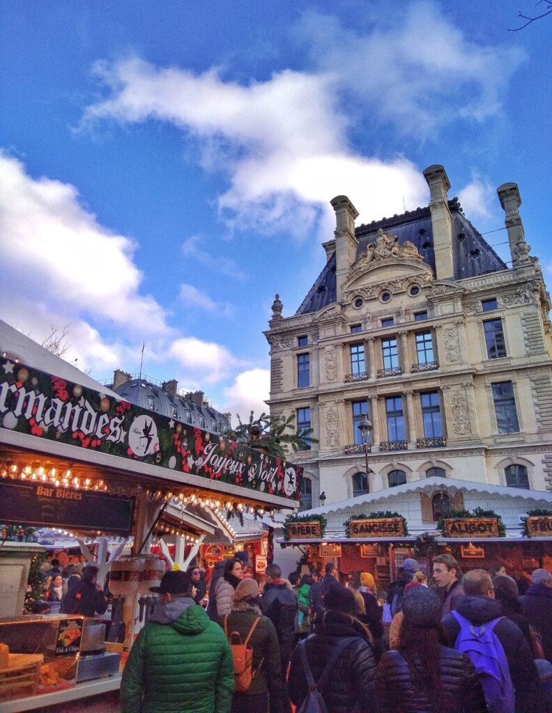 Christmas Market at the Tuileries gardens in Paris. Crowds of people in front of the food stalls, and the Louvre building in the background.