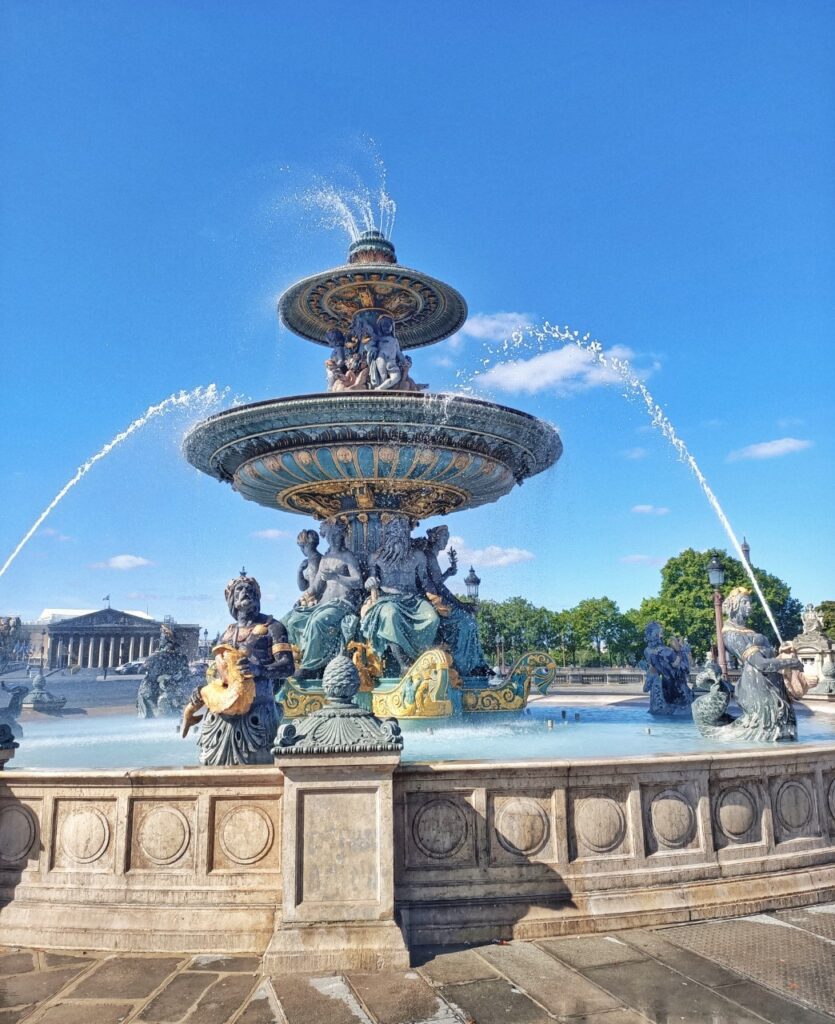 One of the fountains at the Place de la Concorde, in Paris. The fountain is on and we can see water droplets flying around. The sky is blue and we can see the Assemblée Nationale building in the background, looking like a Greek temple.
