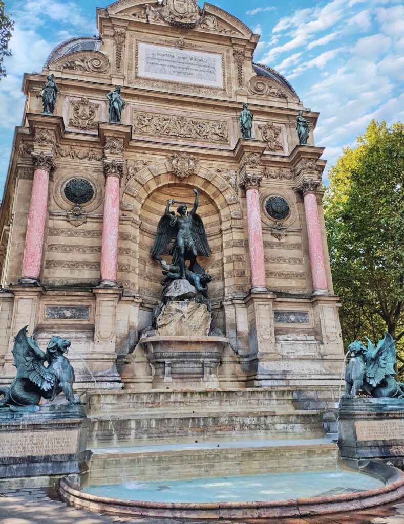 The St Michel fountain in Paris. The water is on a cascading down the fountain. We can see the blue sky and spring trees in the background.