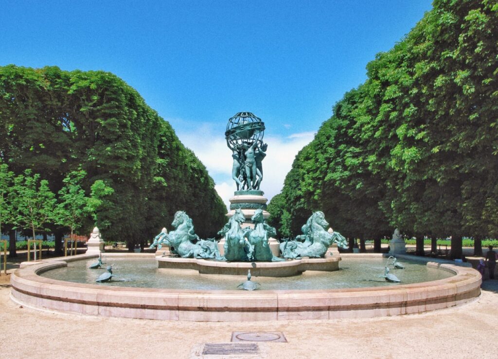 Fountain of the Observatory in Paris. We can see the statue inbetween two rows of trees. The sky is blue in the background.