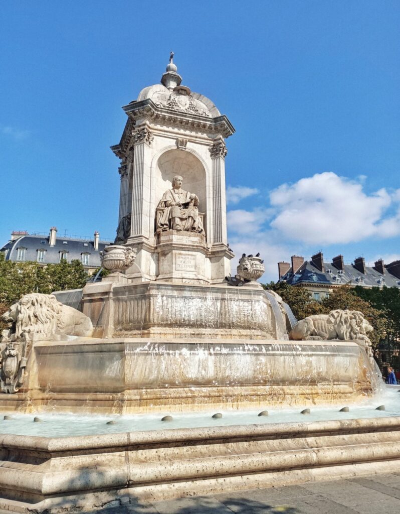 Fontaine St Sulpice in Paris, on a sunny day. There is water running down the fountain and a sculpture at the top. In the background there are typical Parisian buildings.