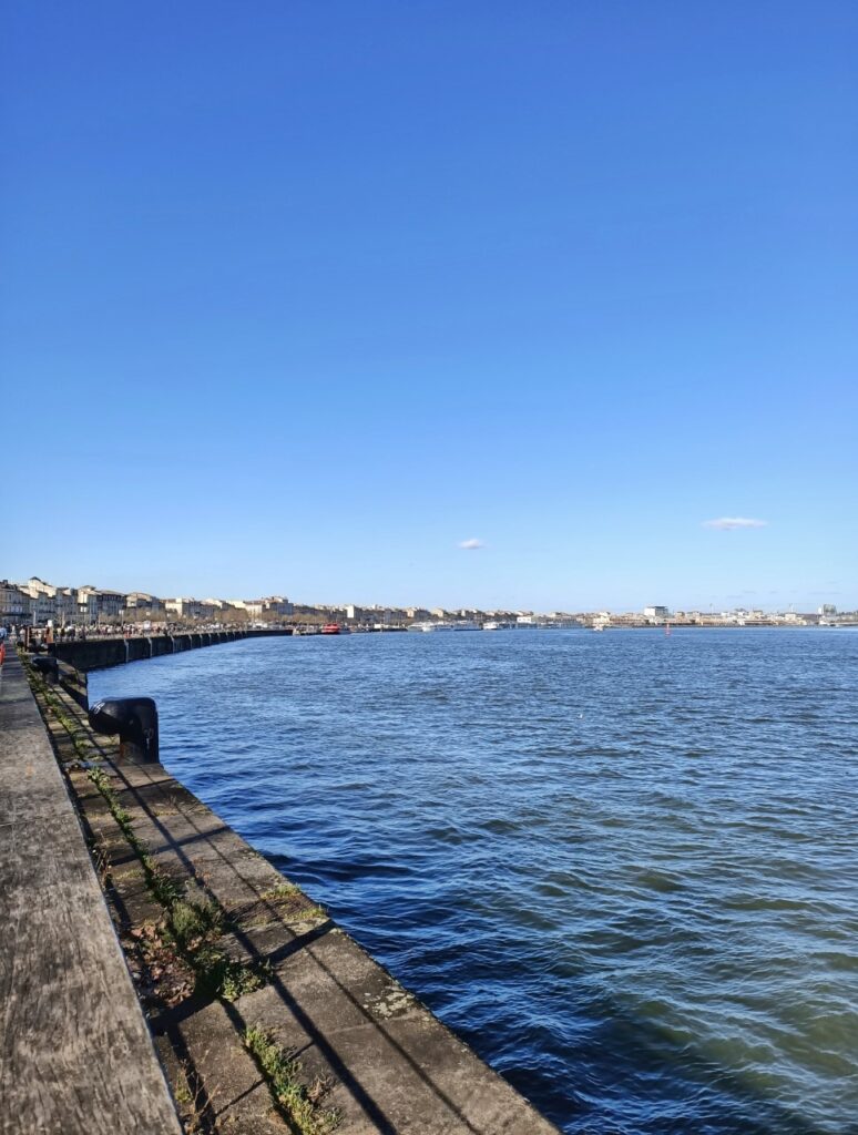 Promenade along the Garonne River in Bordeaux, France. We can see the blue waters of the river and a line of buildings along it.