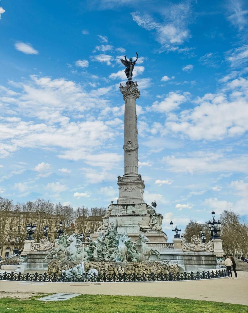 The Monument aux Girondins in Bordeaux, France. The sculptures of the fountain are in the foreground and the tower rises high in the blue sky.
