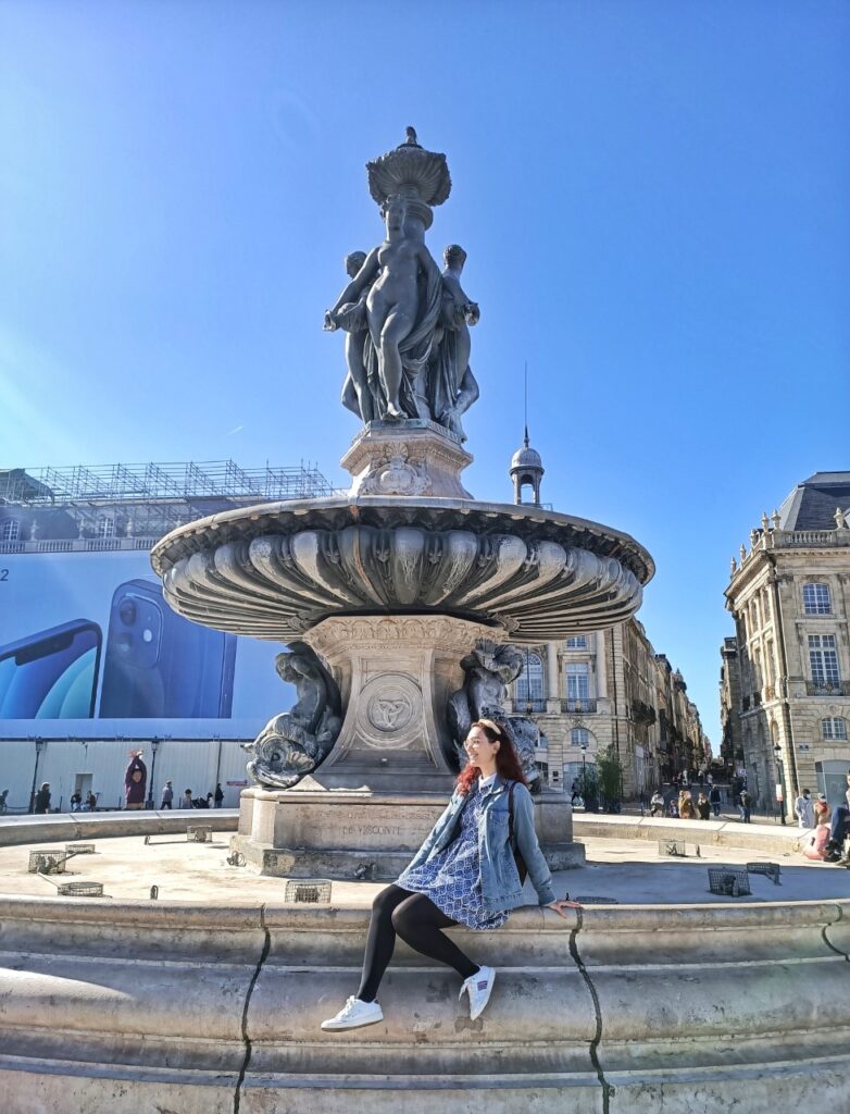 The fountain at Place de la Bourse in Bordeaux, France. I am sitting on the side of the fountain. In the background there are the buildings typical of this area of Bordeaux.