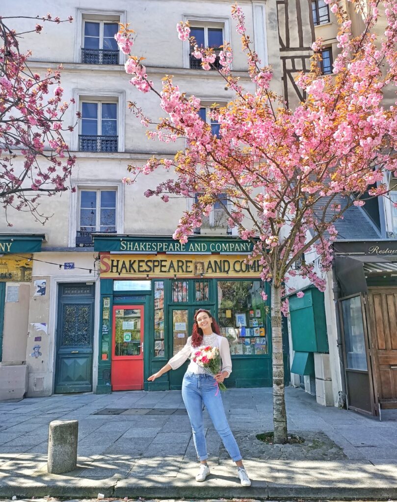 I am standing under a cherry blossom tree, in front of the Shakespeare and Co bookshop in Paris, France. The cherry blossom tree is full of pink flowers and I am holding a pink and red bouquet in my hand.