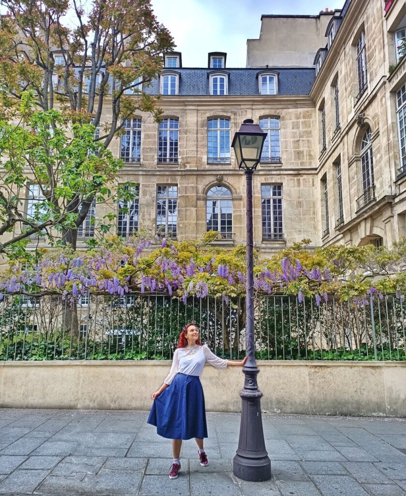 I am standing next to an old-fashioned lamppost in the Rue de Fourcy in Paris. Behind me is a long wisteria plant full of purple flowers.