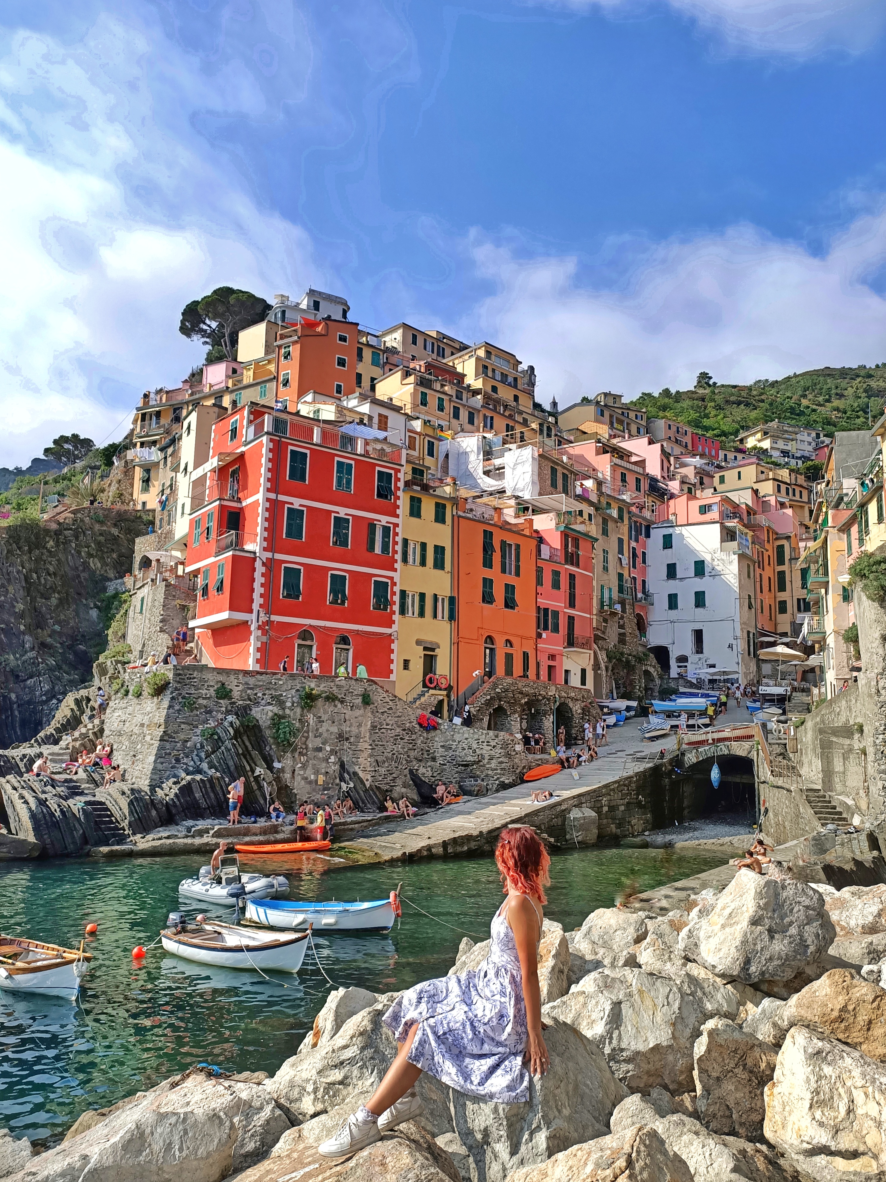 Sunset spot in Riomaggiore, Cinque Terre. I am sitting on the rocks near the water, we can see small fishermen's boats in the background and the typical colourful houses of Riomaggiore.