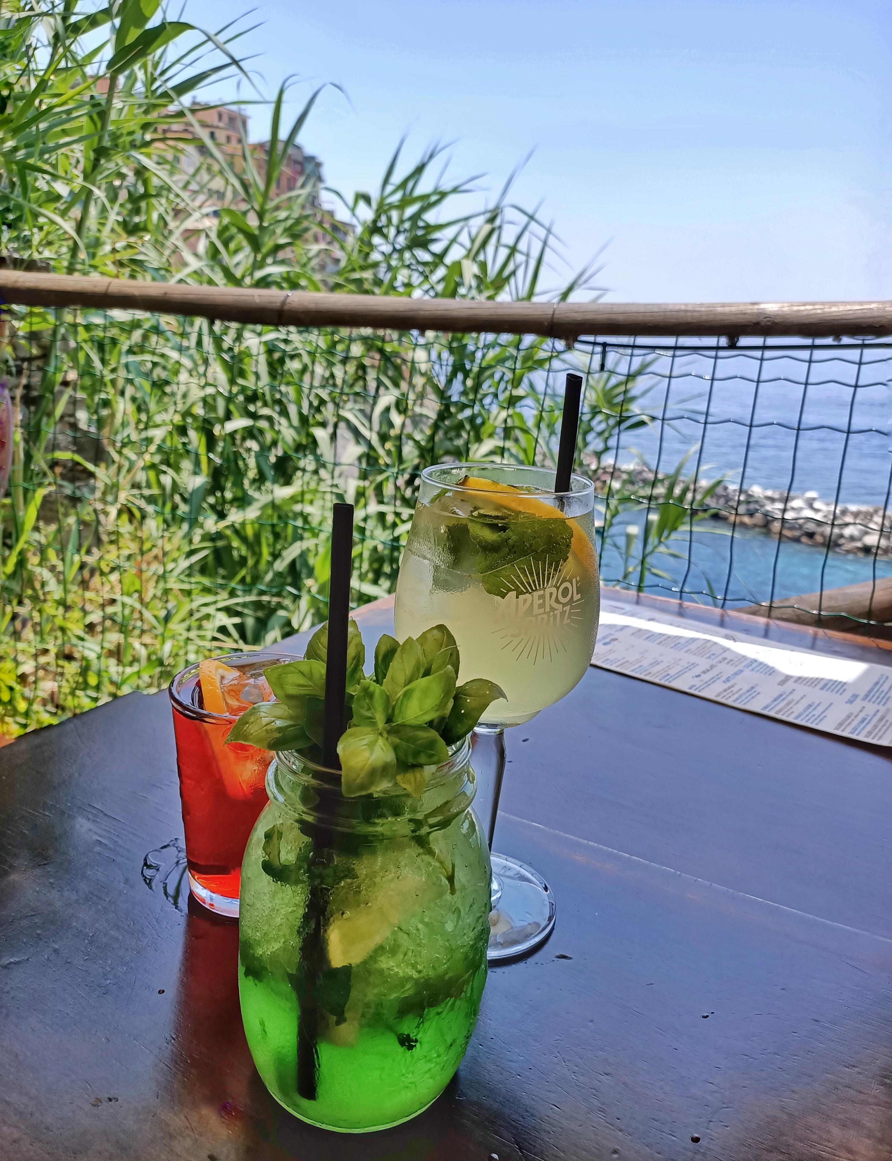 Three cocktails at the Nessun Dorma bar in Manarola, Italy. Their famous cocktail is the Basilito, a basil and mint cocktail. In the background, we can see the sea and the colourful houses of Manarola.