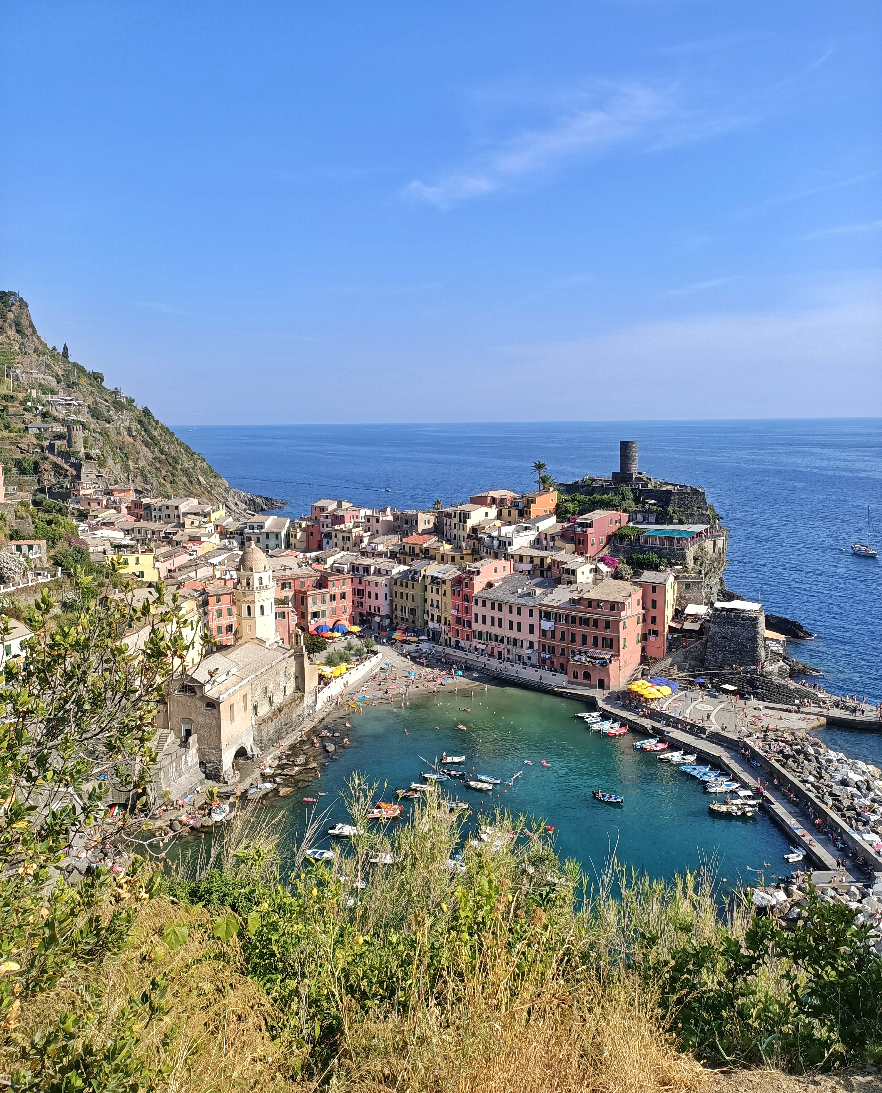 A panoramic view of Vernazza, a village of the Cinque Terre in Italy. We can see the blue water of the sea, the boats, and the colourful houses of the village.