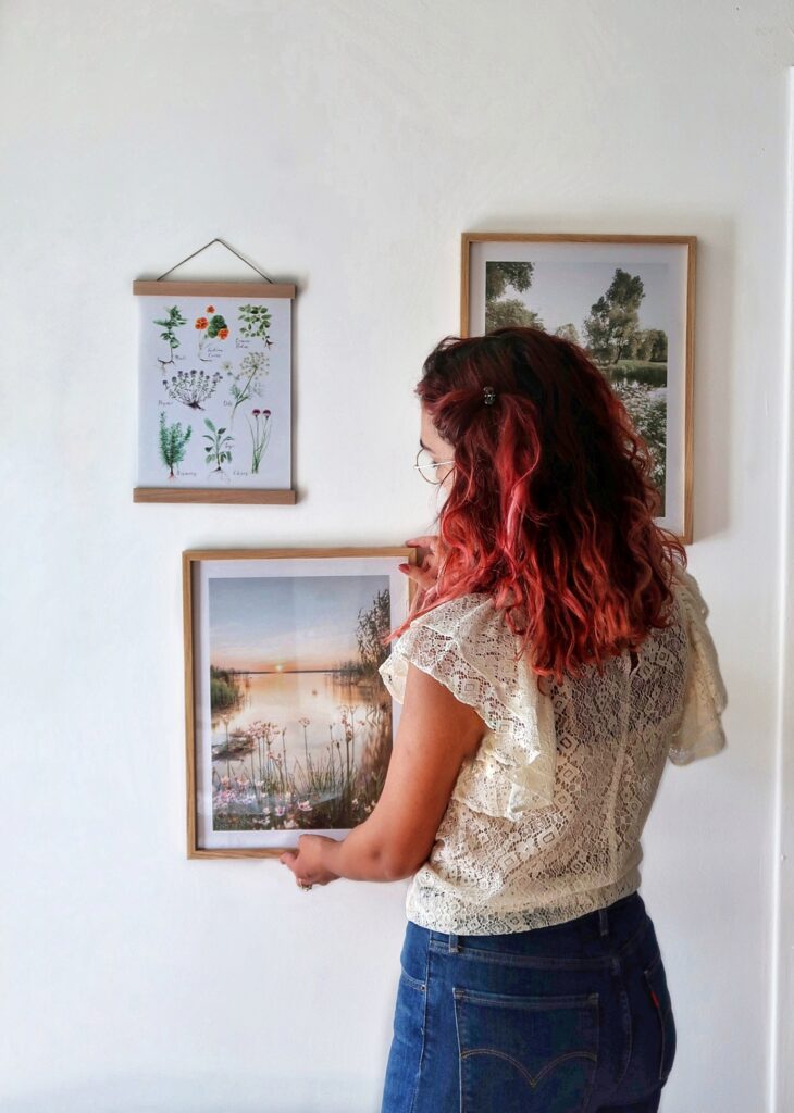 Picture of me facing a white wall in my house. I am hanging a framed poster to complete my wall art gallery. The posters are of nature, landscapes and plants.