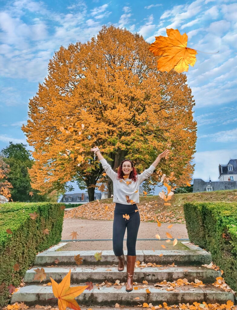 Autumnal photo taken in Blois, France. I am standing on some stairs, throwing orange leaves in the air. I am smiling. Behind me is a big tree full of orange leaves.