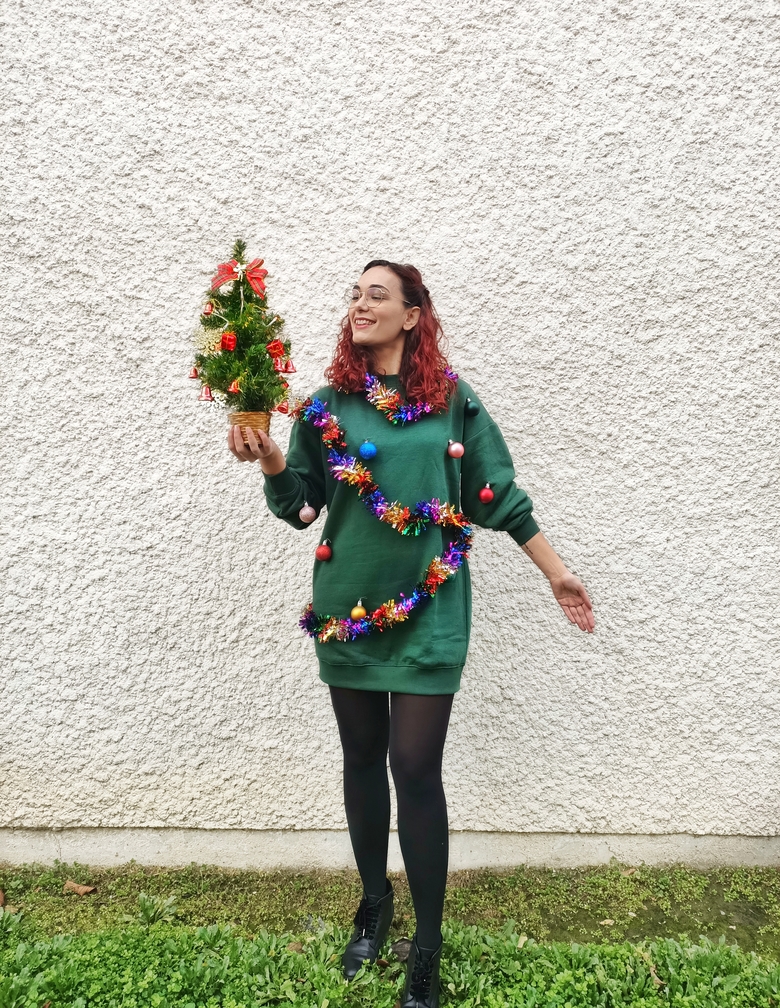 Picture of me wearing a Christmas tree dress with tinsel and baubles. I am holding a small Christmas tree in my hand.