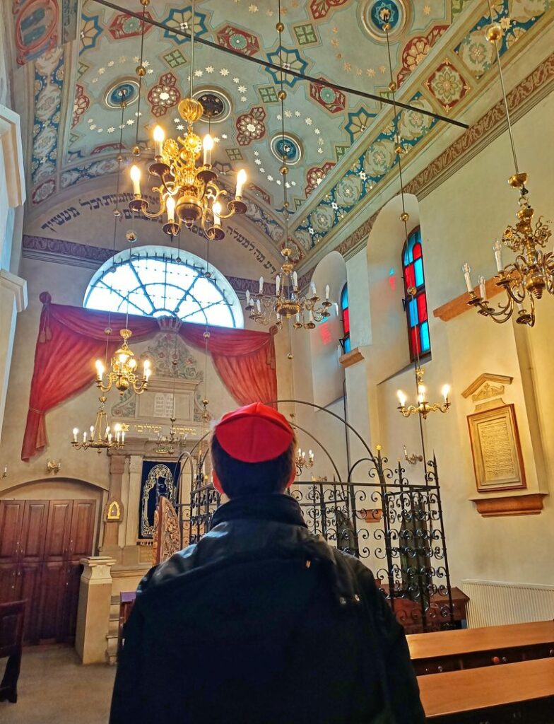 Man standing in the Remuh synagogue in Kazimierz, the Jewish Quarter of Krakow, Poland. There are chandeliers hanging from the ceiling and ornate decorations on the ceiling.