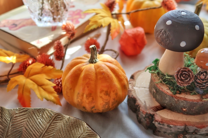 Autumn home decor showing a small orange pumpkin, wooden pieces and a wooden decorative mushroom. In the background, there is a garland of orange autumn leaves and a book.