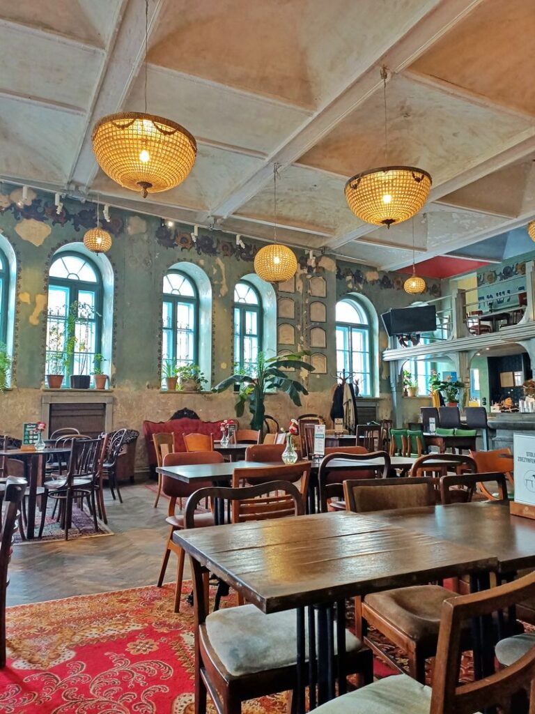 The interior of Hevre, a café in Kazimierez, Krakow, Poland. The wooden furniture and art deco chandeliers give a cosy vibe to the café. On the walls, we can see the remnants of paintings, from the old Jewish praying house the building used to be.