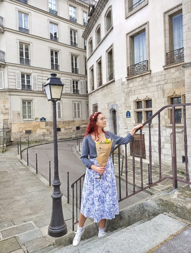 I am standing on stairs with a bouquet of mimosa in my hands. I am in Paris, France. Next to me is a lamppost. I am wearing a white and blue midi dress and a grey cardigan.