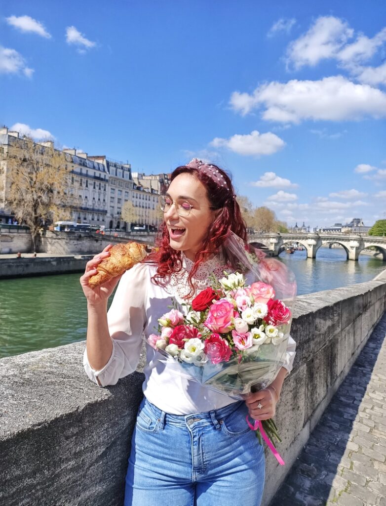 Picture taken in Paris in the spring. I am holding a croissant and a bouquet of red and pink flowers. Behind me is the river Seine and a bridge. There are some typical Parisian buildings on the left side of the bank. The sky is blue.