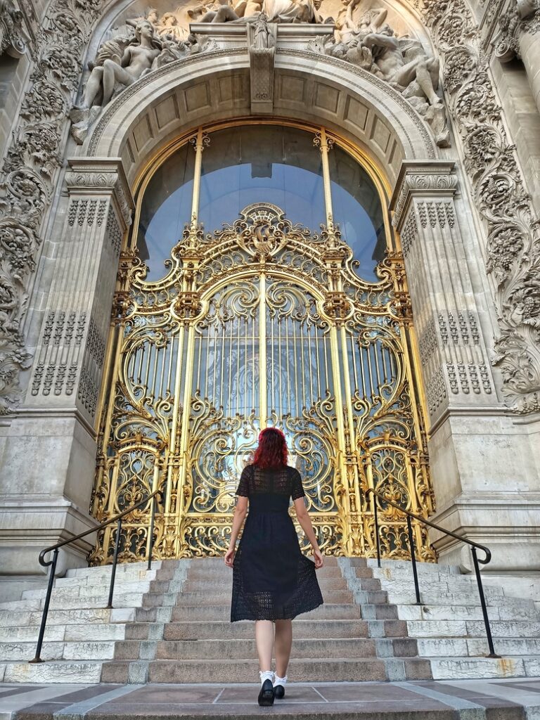 I am standing in front of the imposing golden gate of the Petit Palais museum in Paris, France. The gate is very intricate. I am wearing a black midi dress.