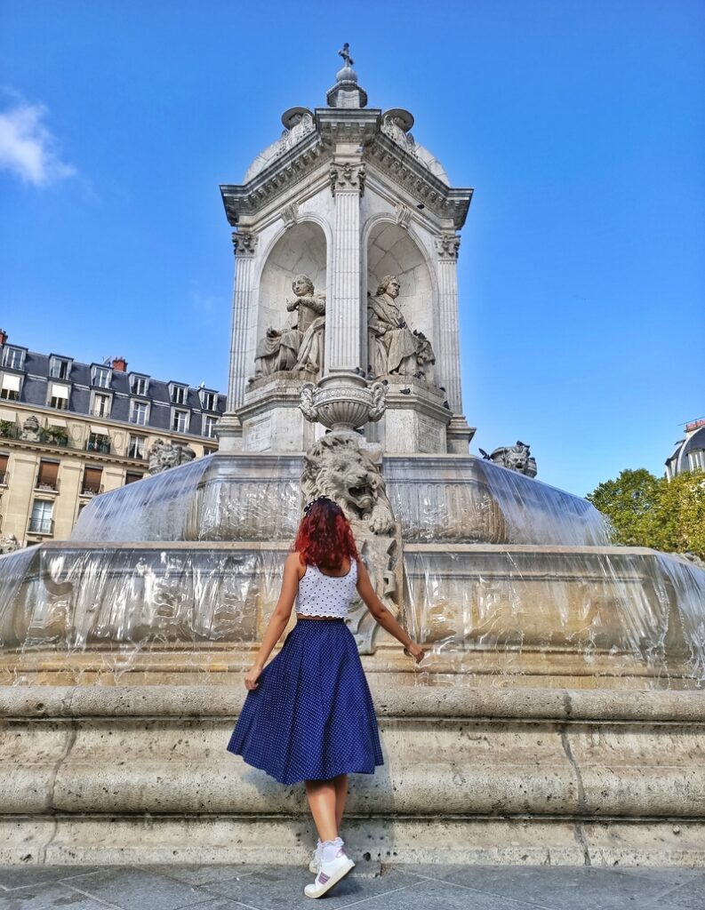 I am standing in front of the Saint Sulpice fountain  in Paris, France. I am wearing a blue midi skirt and crop top, it is the summer and the sky is blue. The water is running in the fountain.