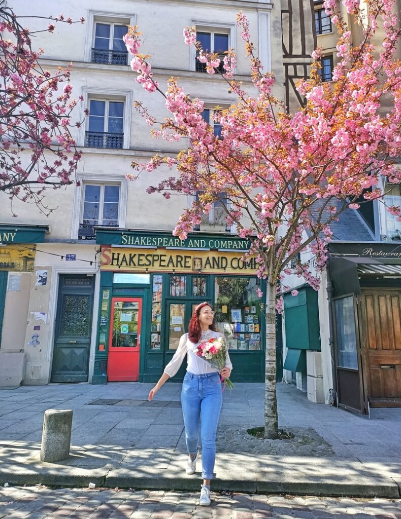 I am holding a bouquet of pink and red flowers and standing in front of the Shakespeare and Co bookshop in Paris, France. There is a pink cherry blossom tree in full bloom next to me.