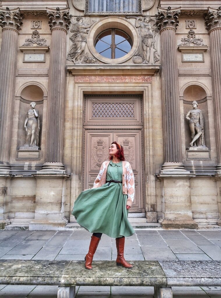 I am standing on a bench in front of the Louvre Museum building in Paris, France. I am wearing a green cottagecore dress and a chequered knitted cardigan. My skirt is flowing in the wind. Behind me are some statues and columns of neoclassical architecture.
