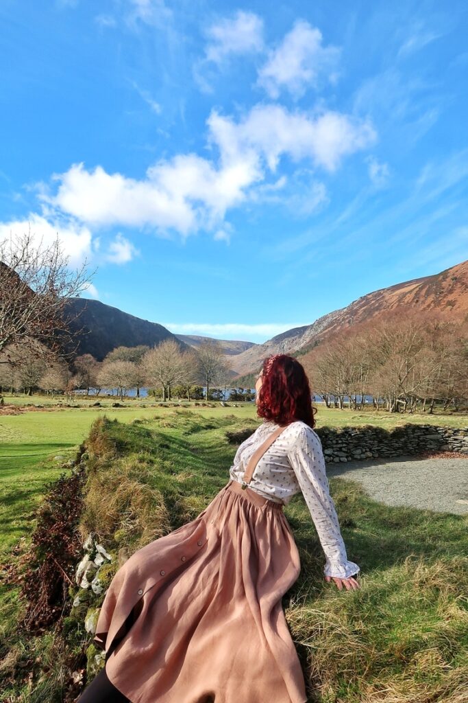 I am looking at the Glendalough mountains and lake in Wicklow, near Dublin, Ireland. There is a lot of greenery and the sky is blue.