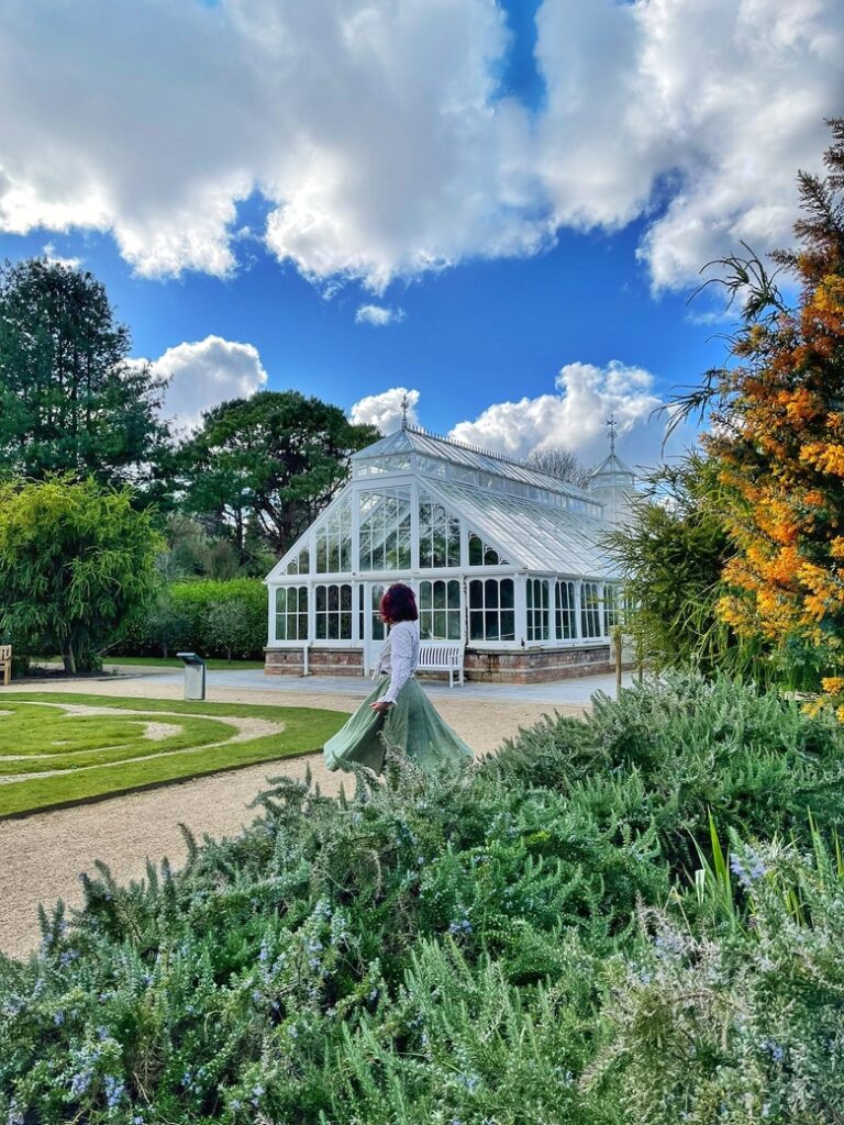 I am standing in the botanical gardens at Malahide Castle, near Dublin, Ireland. I am twirling and there is a Victorian greenhouse behind me. There are yellow and purple flowers around me.
