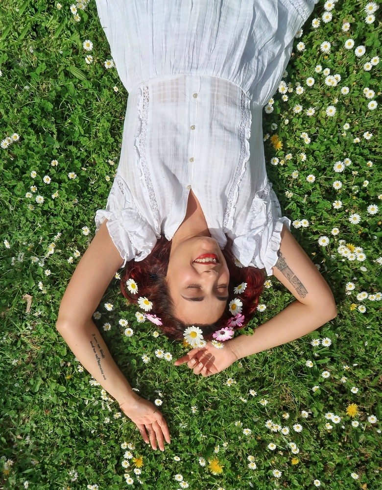 Picture of a woman laying on the grass, surrounded by daisies. She is wearing a white dress and is smiling. She feels peace and she has learned to slow down and improve her life.