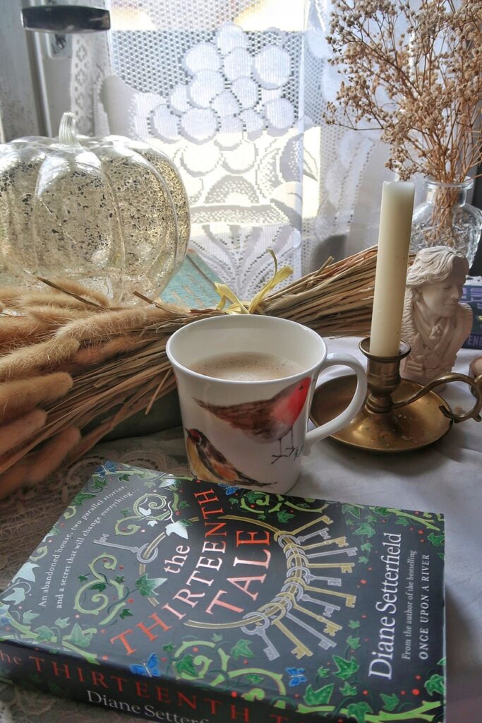 A pretty book (The Thirteenth Tale by Diane Setterfield) is sitting on a desk, with a cup of coffee and a candle holder beside it.