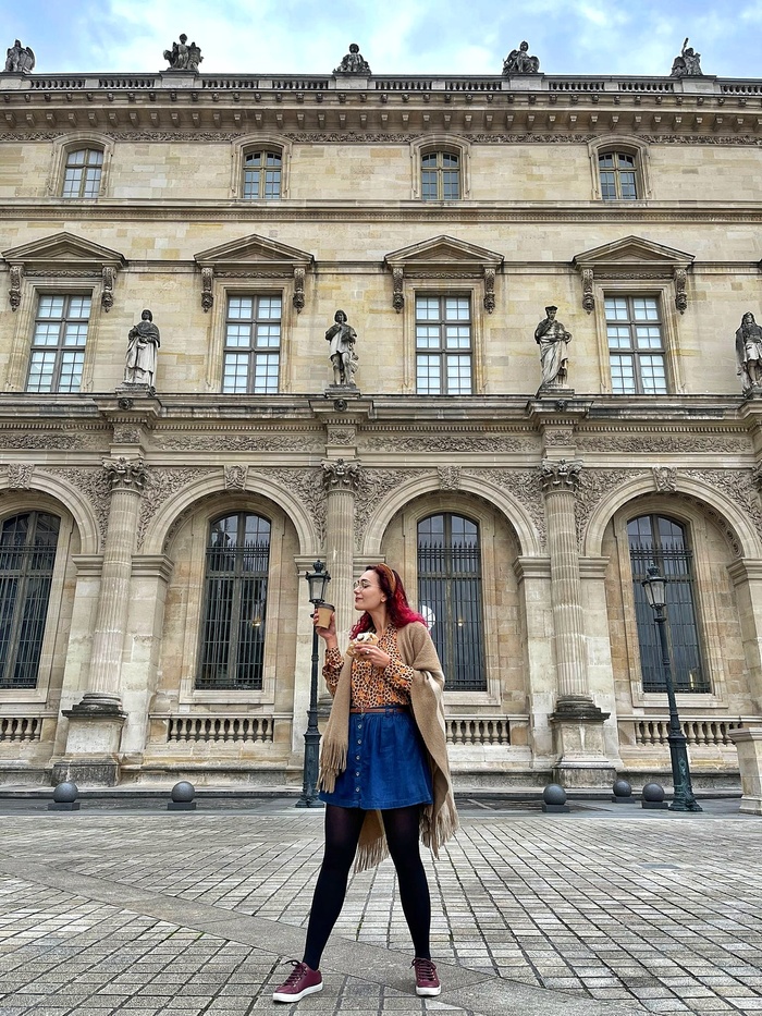 Julia is standing in front of the Louvre in Paris. It is autumn. She is drinking a pumpkin spice latte and eating a cinnamon bun.