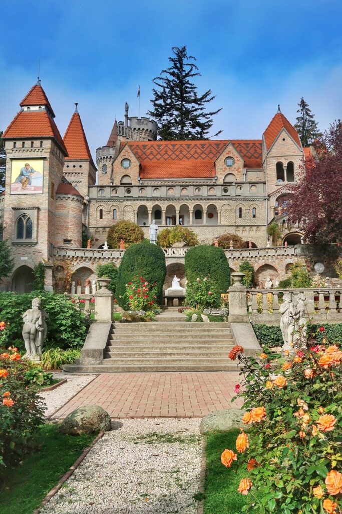 The picture depicts the facade of Bory Castle, in Hungary. The fairytale castle has many towers, a red tiled roof and terraces with sculptures. There are rose bushes in the front garden.