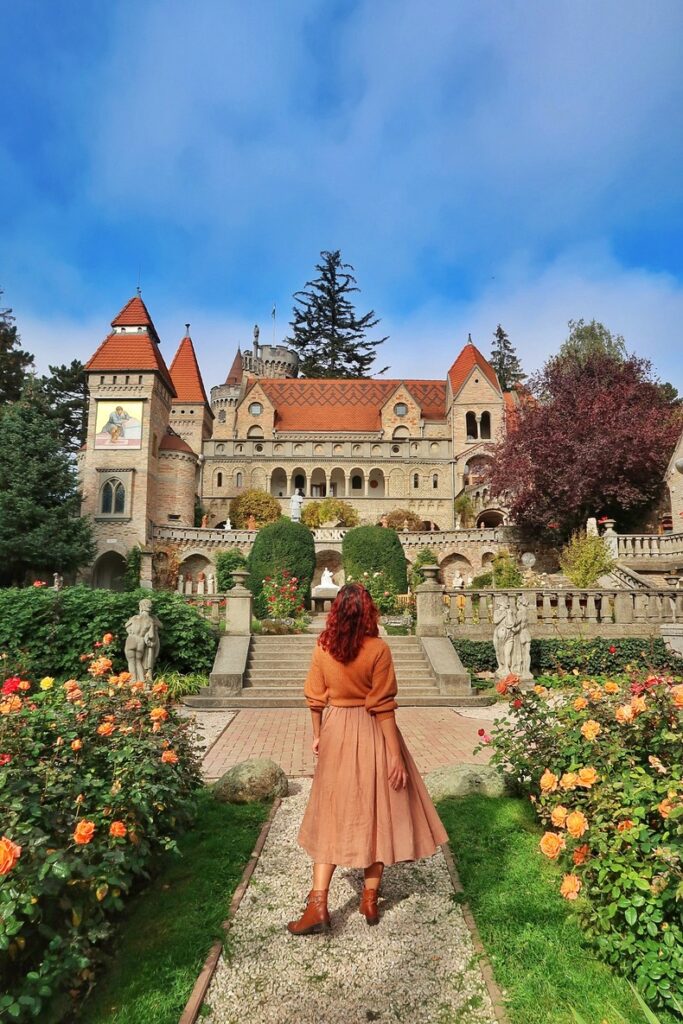 A girl (julia-speaks.com) is standing in front of Bory Castle. She is facing the beautiful Hungarian castle. There is a rose garden around her. The castle is made of several fairytale towers and terraces. 