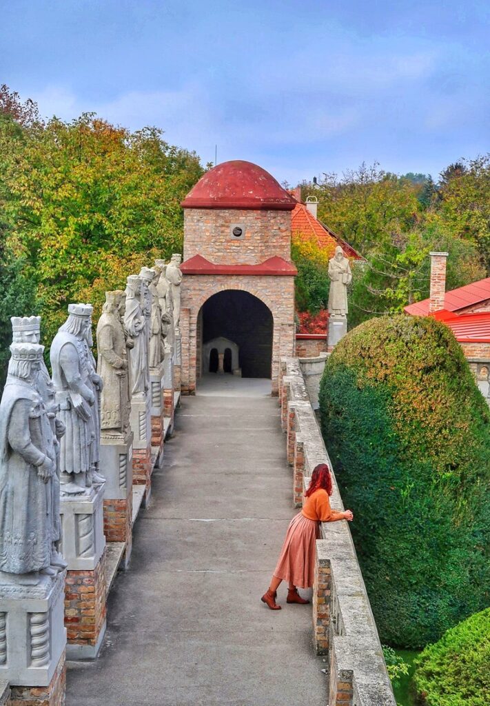 A view from the courtyard tower of Bory Castle in Hungary. Julia (julia-speaks.com) is standing on the edge, looking in the distance. There are tall statues on the other side of the courtyard. There are autumn trees in the background.
