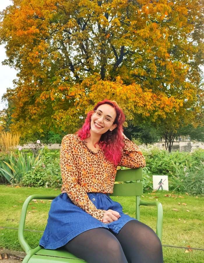 Julia is sitting on a green chair at the jardin des Tuileries in Paris. She is wearing an autumnal leopard print shirt and a denim skirt. There is an orange tree behind her.