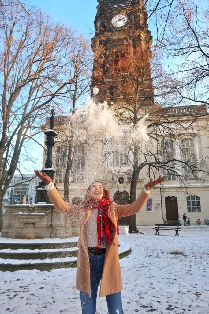 Julia is standing in front of a church in Dresden, Germany. There is snow on the ground, it is the winter. Julia is throwing snow in the air and smiling.
