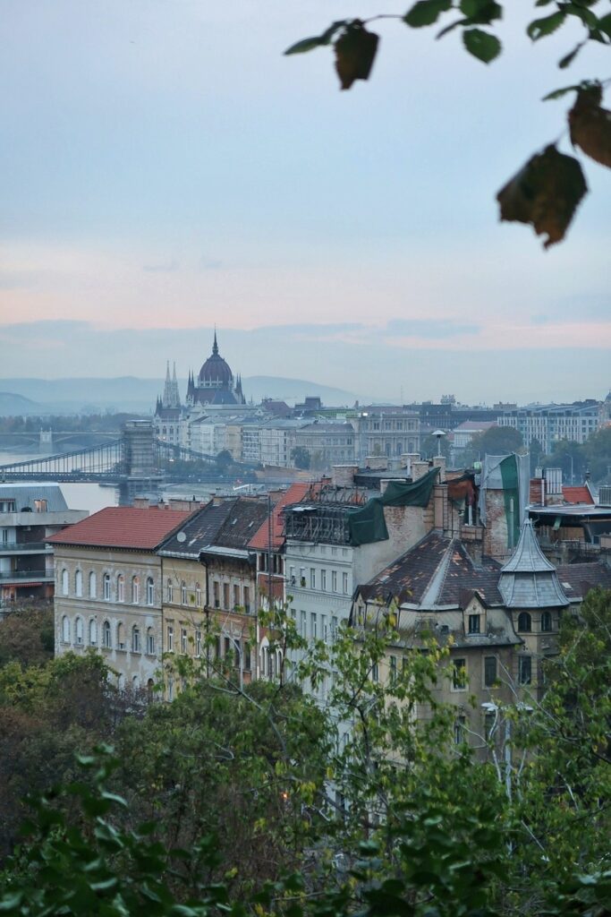 A cloudy sunset view of Budapest from Gellert Hill. We can see the Parliament building and some typical Budapest buildings. The sky is grey and pink.