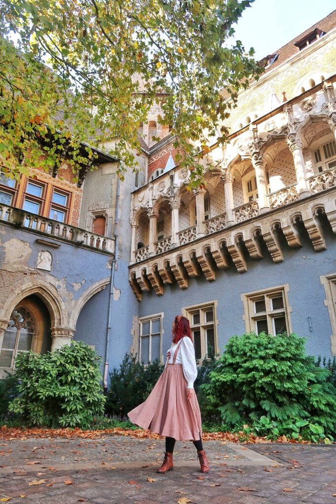 Julia standing in front of a courtyard at Vajdahunyad castle, looking at the Gothic facade. The facade is blue, and there is a whimsical row of columns on a balcony.